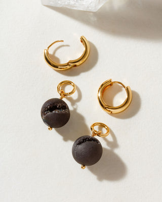A set of gold-colored hoop earrings with one featuring a dark druzy sphere bead charm attached to it, displayed on a white surface.