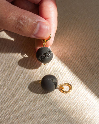 A set of gold-colored hoop earrings with one featuring a dark druzy sphere bead charm attached to it, displayed on a textured surface.