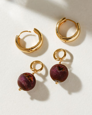 A set of gold-colored hoop earrings with one featuring a colorful druzy sphere bead charm attached to it, displayed on a white surface.