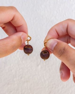 A set of gold-colored hoop earrings with one featuring a colorful druzy sphere bead charm attached to it, displayed on a white surface.