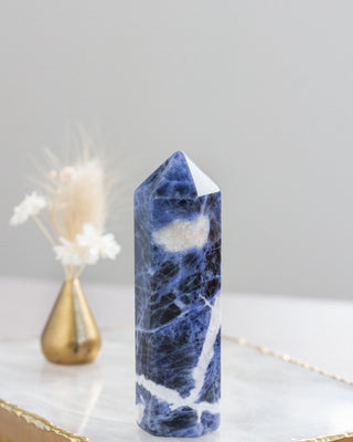 A sodalite gemstone tower with deep blue hues and natural marbled patterns, placed on a white surface with a decorative item in the background.
