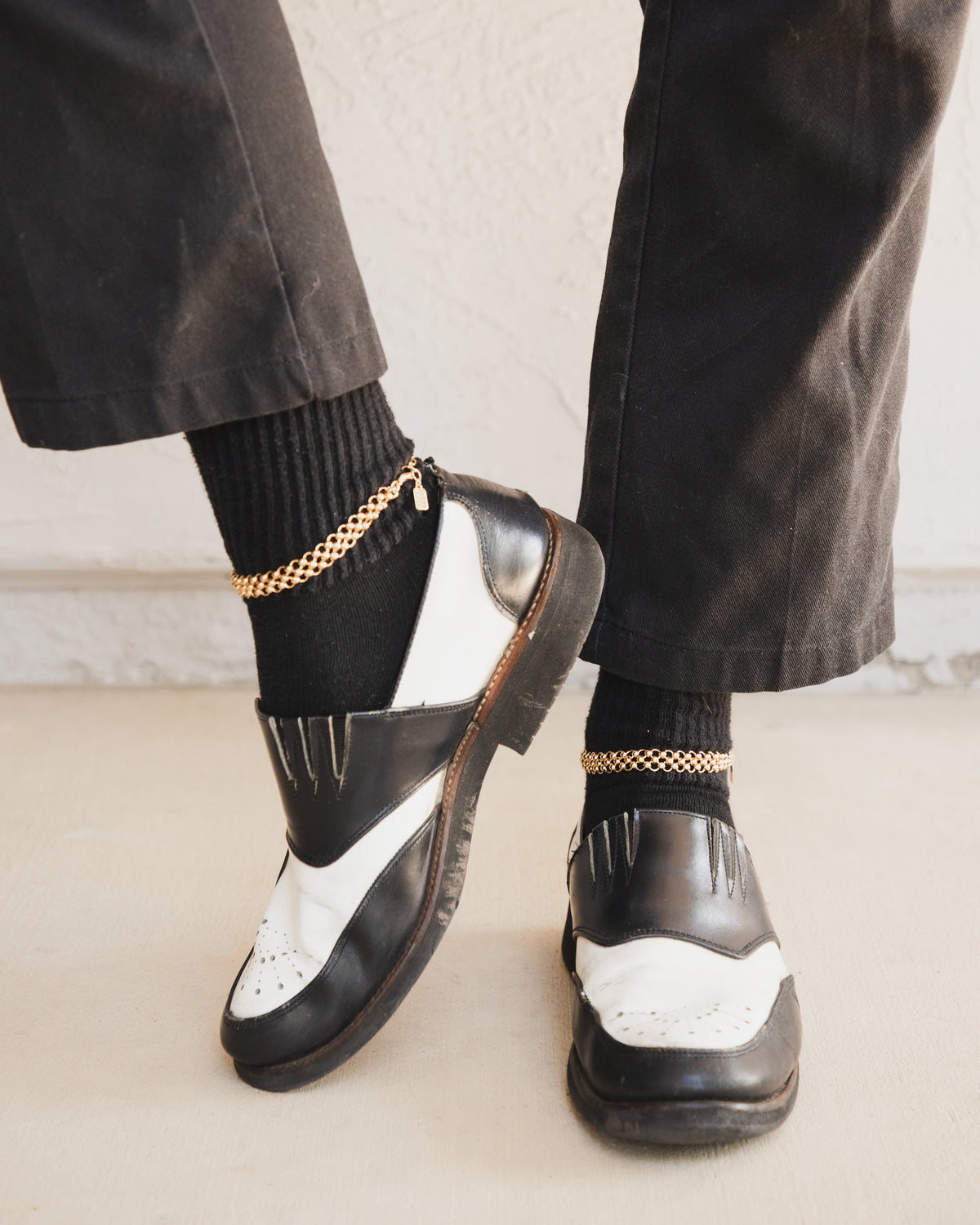 A model is wearing a gold Chain Maille Anklet over black socks with black and white shoes.