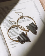 Supernova Earrings in Black, featuring gold hoops with raw crystals, displayed on an open book.