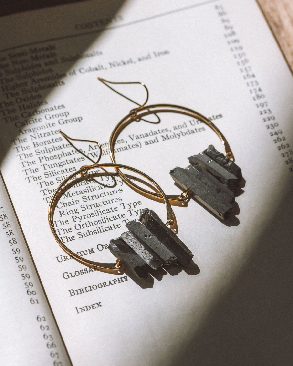 Supernova Earrings in Black, featuring gold hoops with raw crystals, displayed on an open book.