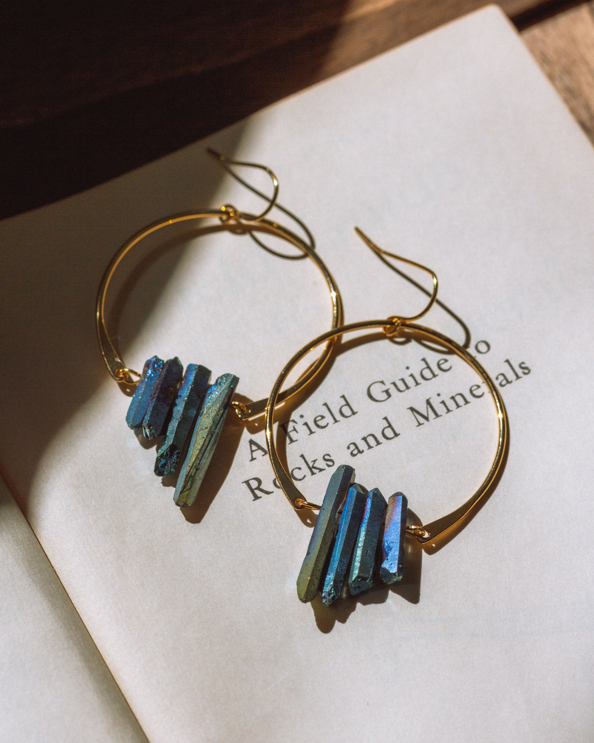 Supernova Earrings in Gold, with a cluster of multicolored metallic blue quartz crystals on a book.