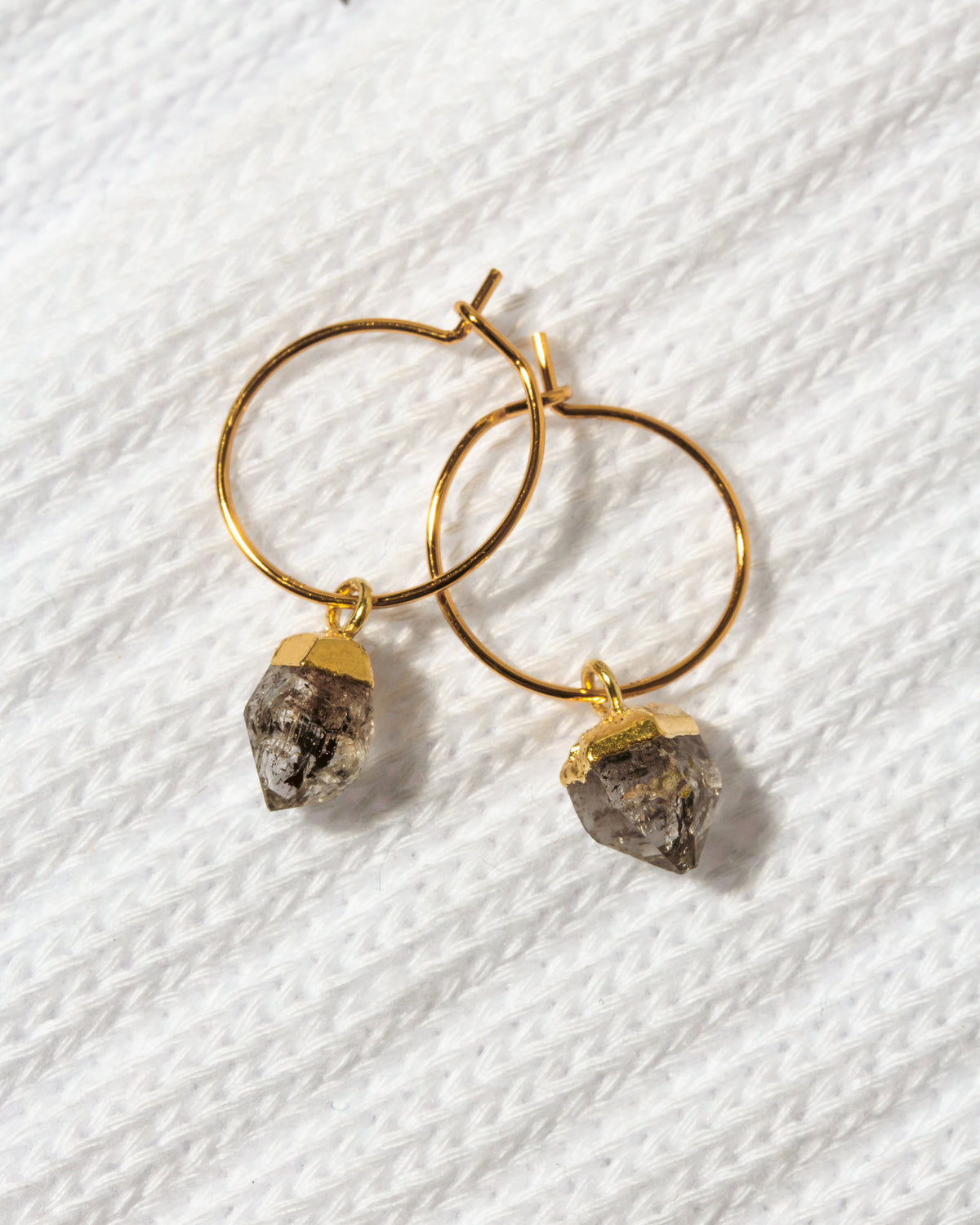 A pair of gold hoop earrings with dangling raw, rough-textured gray Herkimer quartz stones.