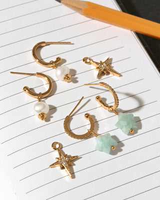 A set of gold-toned dangle earrings arranged on lined paper next to a pencil. The earrings include 2 pairs of hoops with white pearl beads, textured CZ hoops with aquamarine star stone drops, and starburst charms with clear rhinestones.