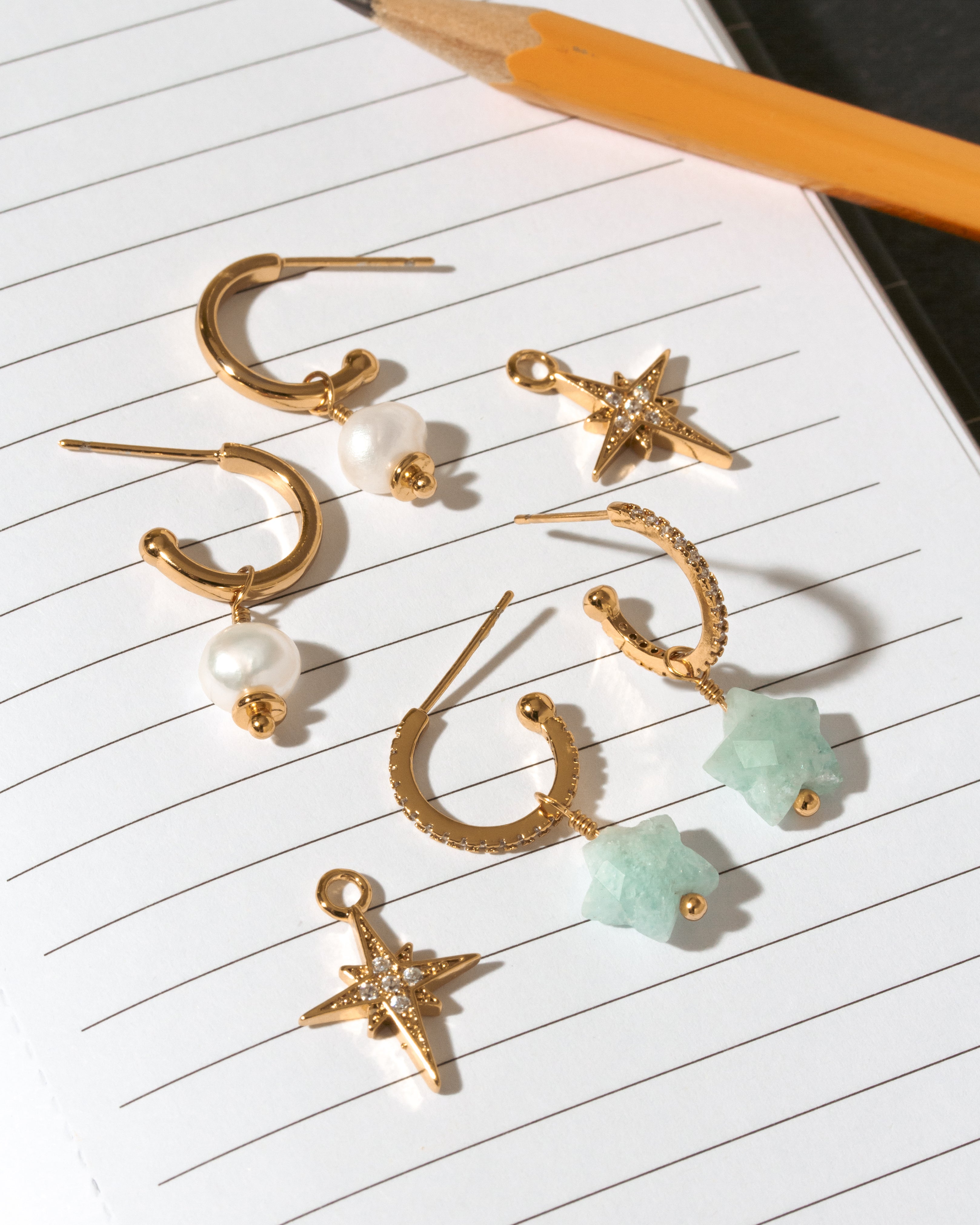 A set of gold-toned dangle earrings arranged on lined paper next to a pencil. The earrings include 2 pairs of hoops with white pearl beads, textured CZ hoops with aquamarine star stone drops, and starburst charms with clear rhinestones.