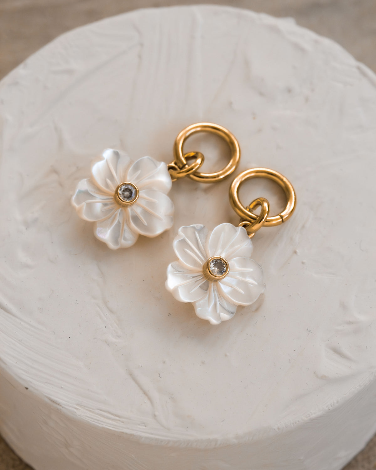 Two earring charms with mother-of-pearl flower design with clear gemstone, on gold hoops.