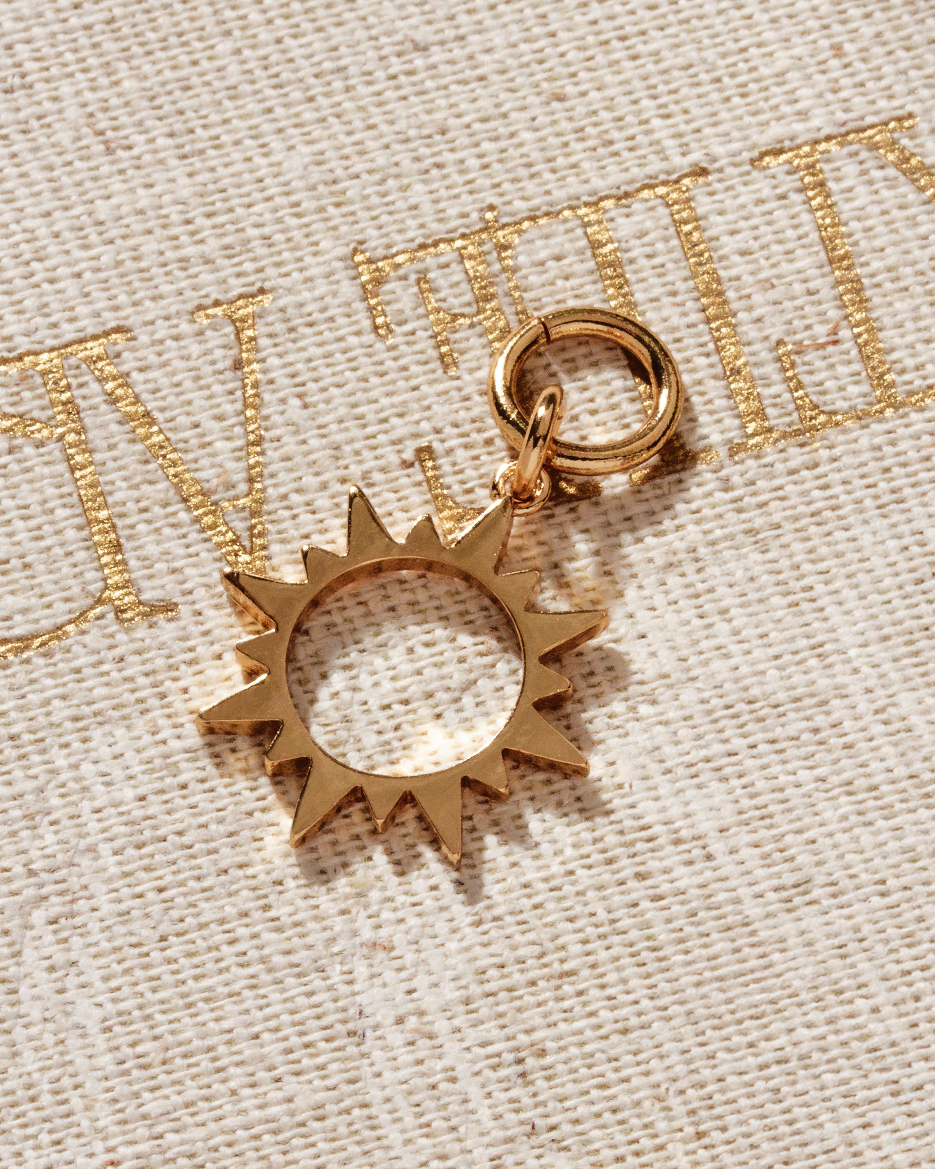 Gold-colored hoop earring charm in the shape of a sun, placed on a textured fabric surface.