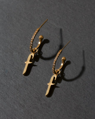 Gold earrings with 'f' charms on a dark gray background