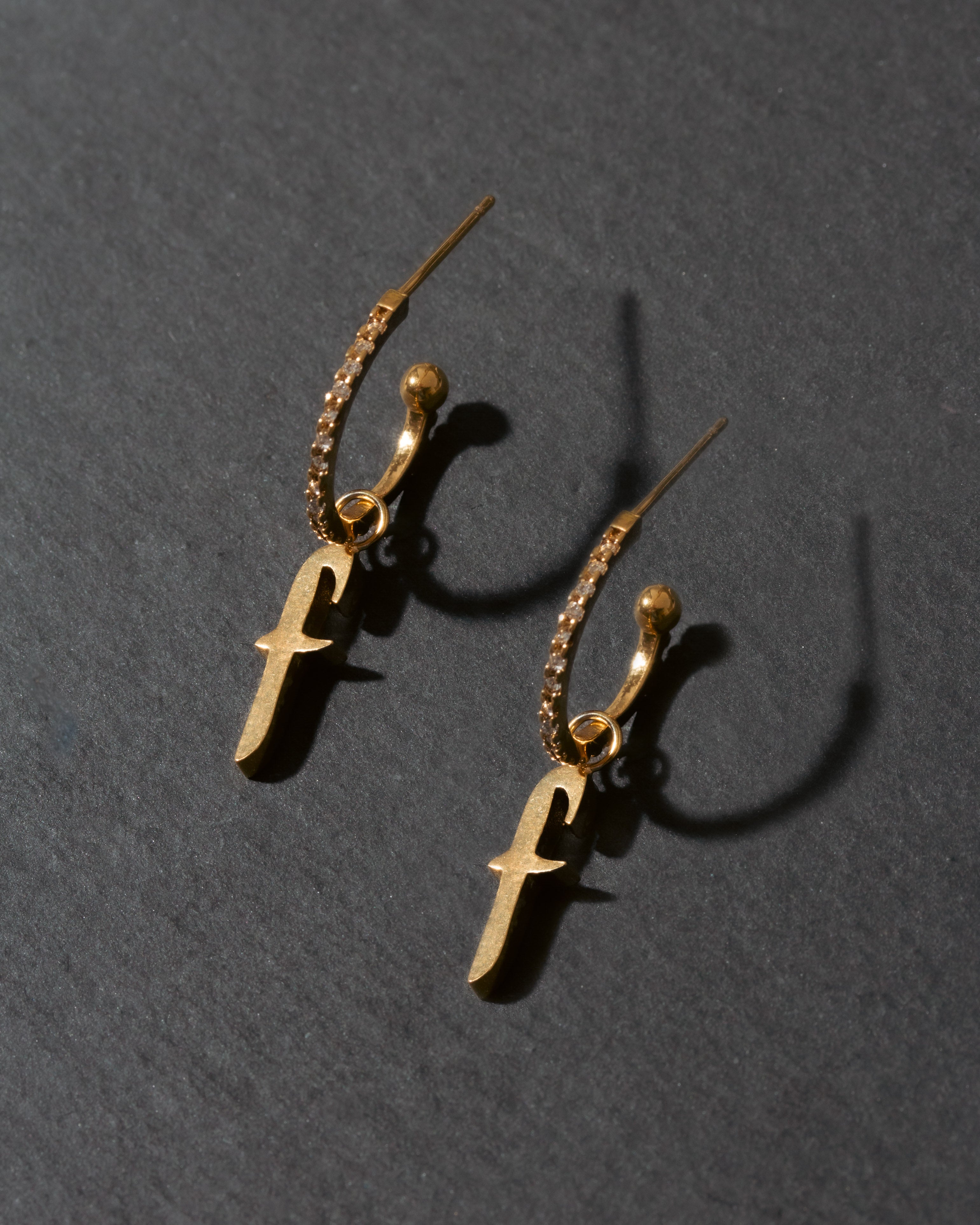 Gold earrings with 'f' charms on a dark gray background
