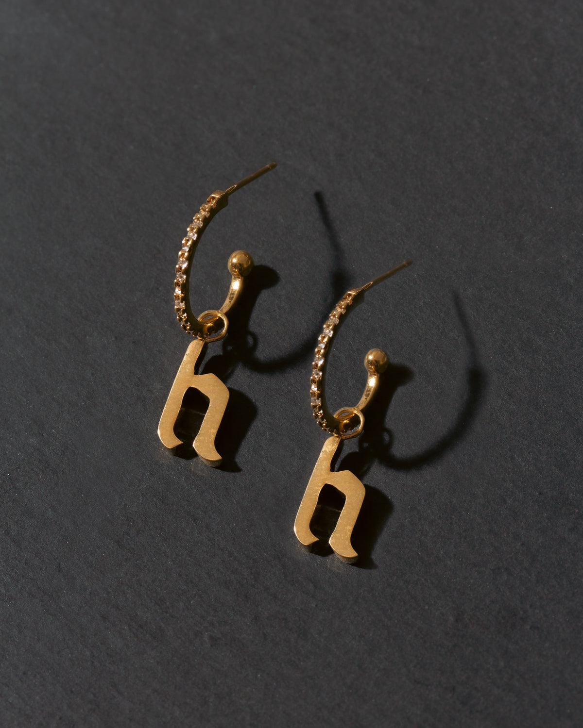 Gold earrings with 'h' charms on a dark gray background