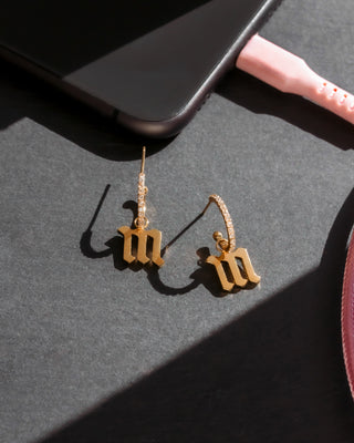 Gold earrings with 'm' charms on a dark surface