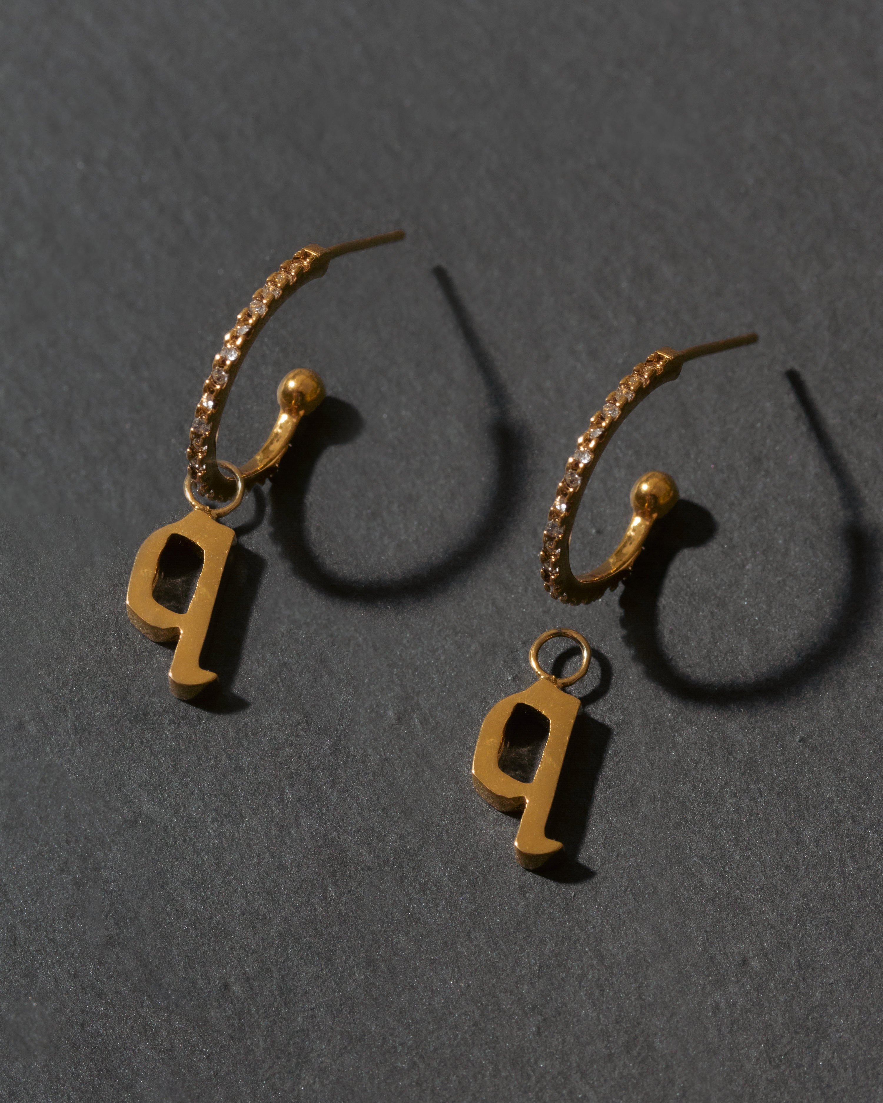 Gold hoop earrings with letter 'q' charms on a dark gray background