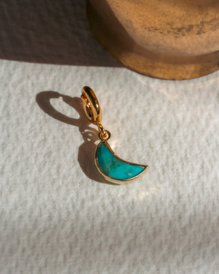 Gold pendant with a turquoise half-moon design on a textured white surface.