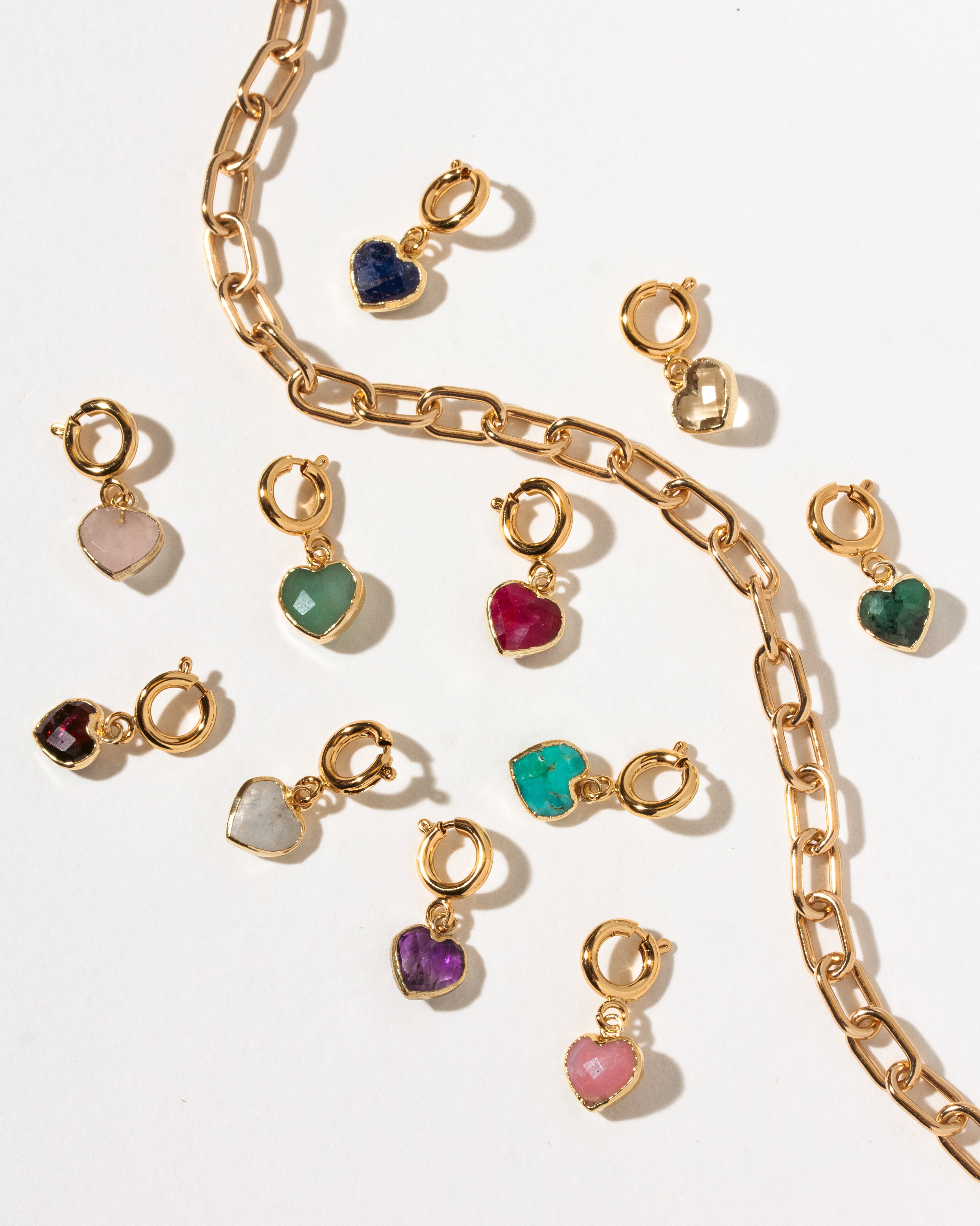 Gold heart-shaped charm clips with various colored gemstones, displayed on a clean white background with a gold paperclip chain.