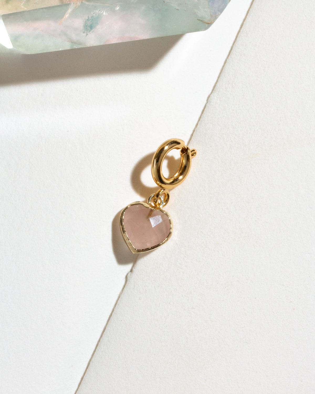 A gold heart-shaped charm with a faceted pale pink rose quartz stone.