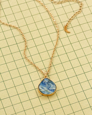 A gold chain necklace with a teardrop-shaped blue and white marbled kyanite pendant, displayed on a grid paper background.
