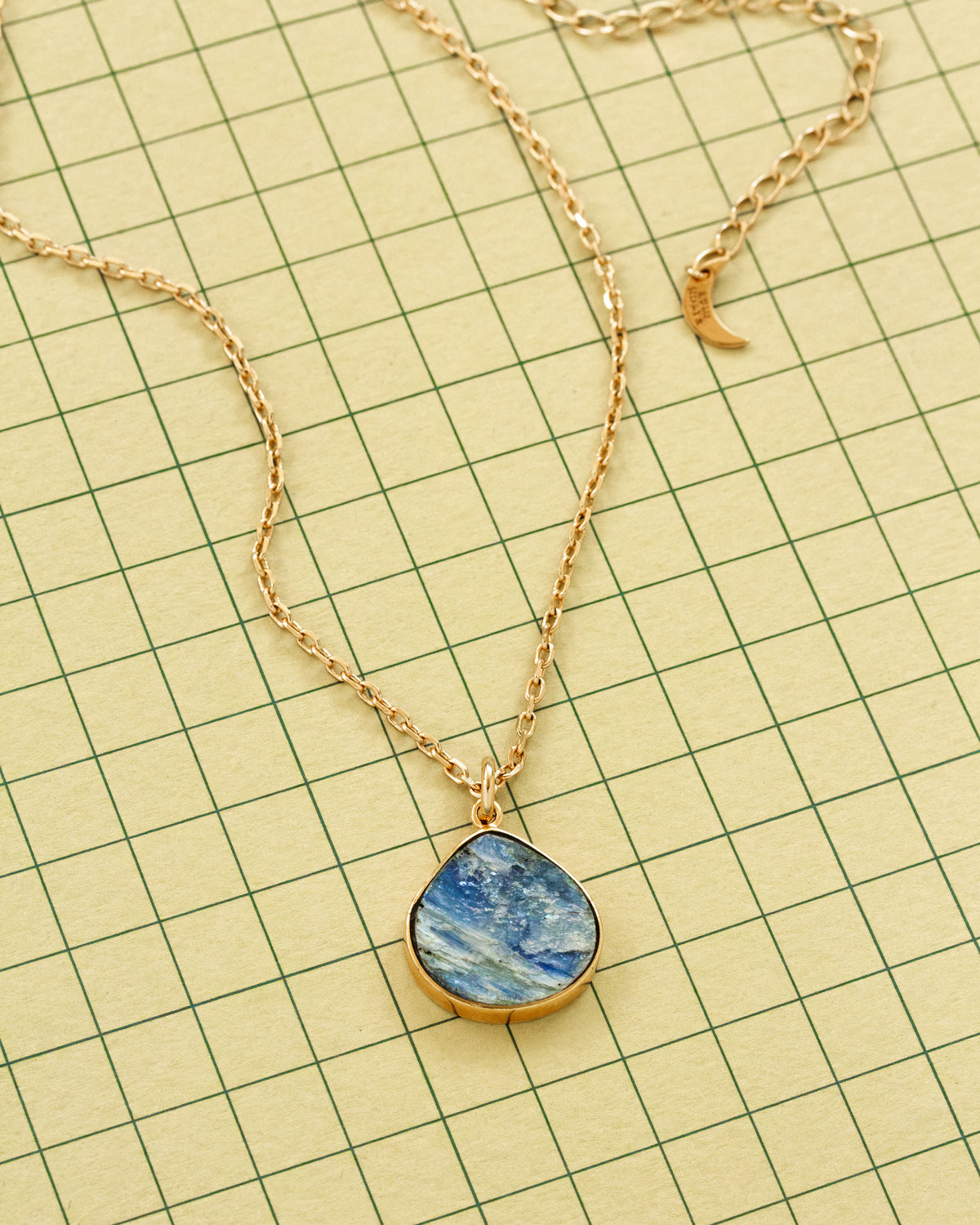 A gold chain necklace with a teardrop-shaped blue and white marbled kyanite pendant, displayed on a grid paper background.