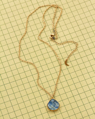 A gold chain necklace with a blue and white marbled kyanite teardrop shaped pendant, laid flat on a yellow grid background.