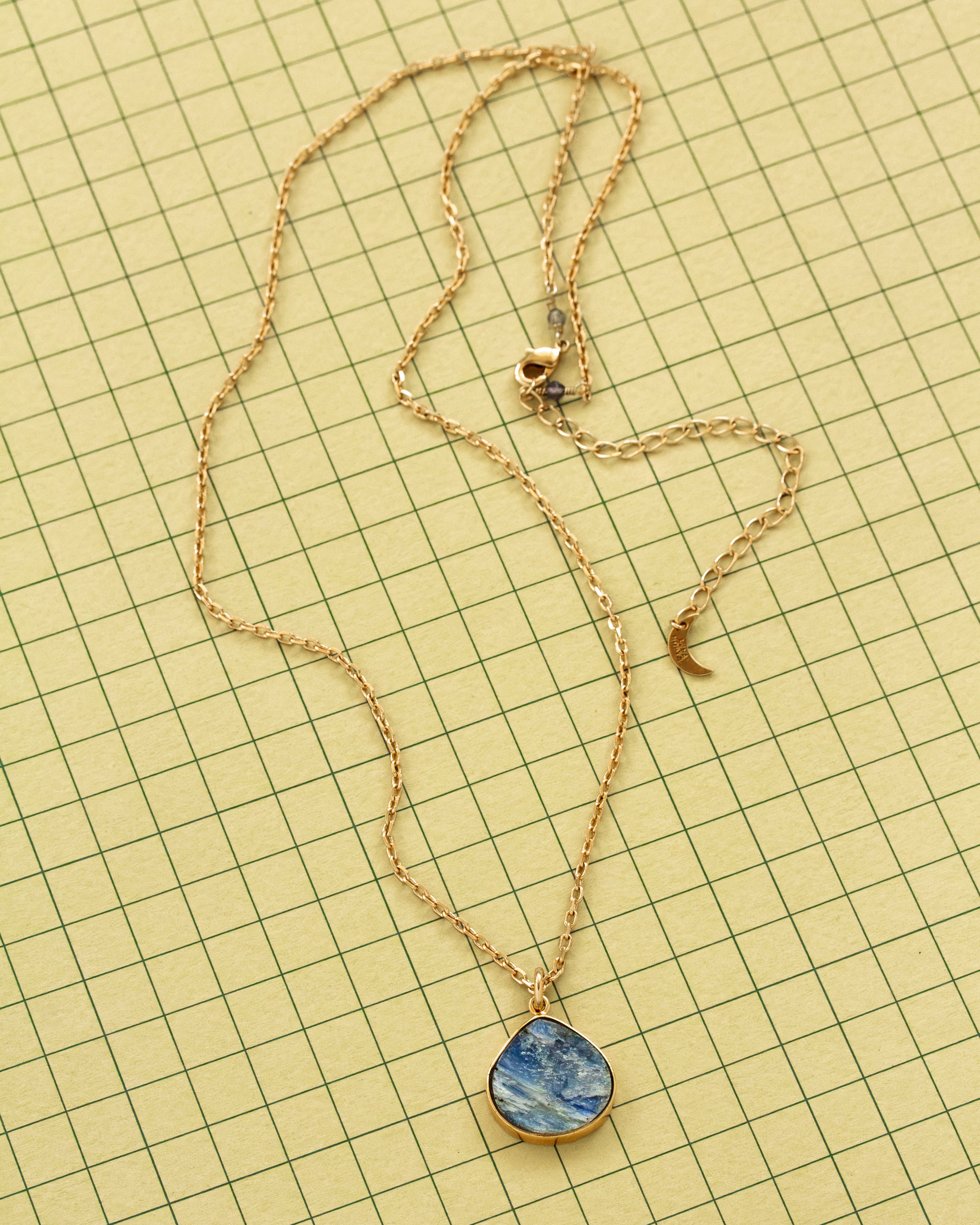 A gold chain necklace with a blue and white marbled kyanite teardrop shaped pendant, laid flat on a yellow grid background.