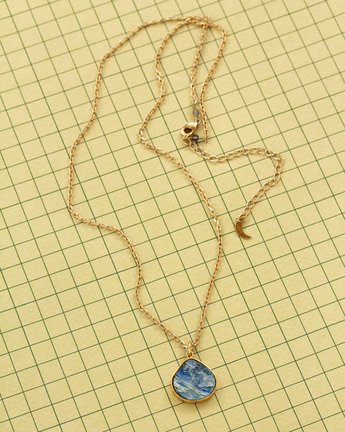 A gold chain necklace with a blue and white marbled kyanite teardrop shaped pendant, laid flat on a yellow grid background.