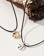 Two delicate cord necklaces with small round hammered pendants, one gold-toned and one silver-toned.