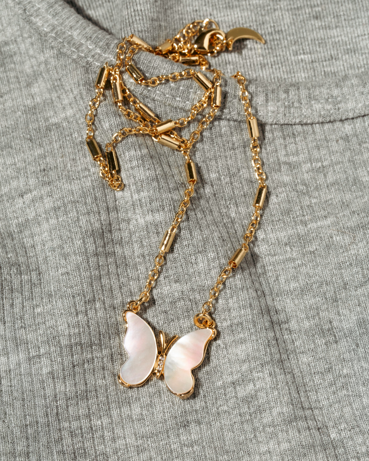 A gold-tone necklace with a mother-of-pearl butterfly pendant, laid flat on a textured gray knit garment.
