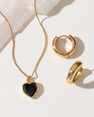 Gold jewelry set featuring a heart-shaped pendant necklace with a black onyx stone inlay and a gold huggie hoop earring.