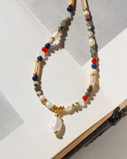 A beaded necklace with a white pearl pendant, featuring white, blue, red, and green beads with gold spacer rings, displayed on an open book.
