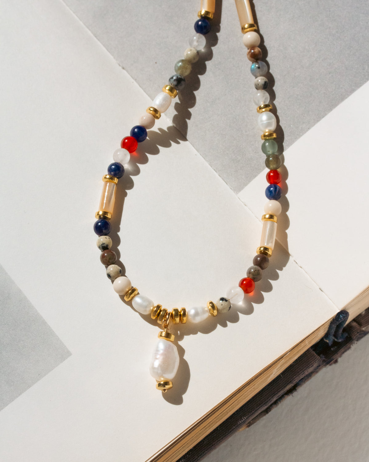 A beaded necklace with a white pearl pendant, featuring white, blue, red, and green beads with gold spacer rings, displayed on an open book.
