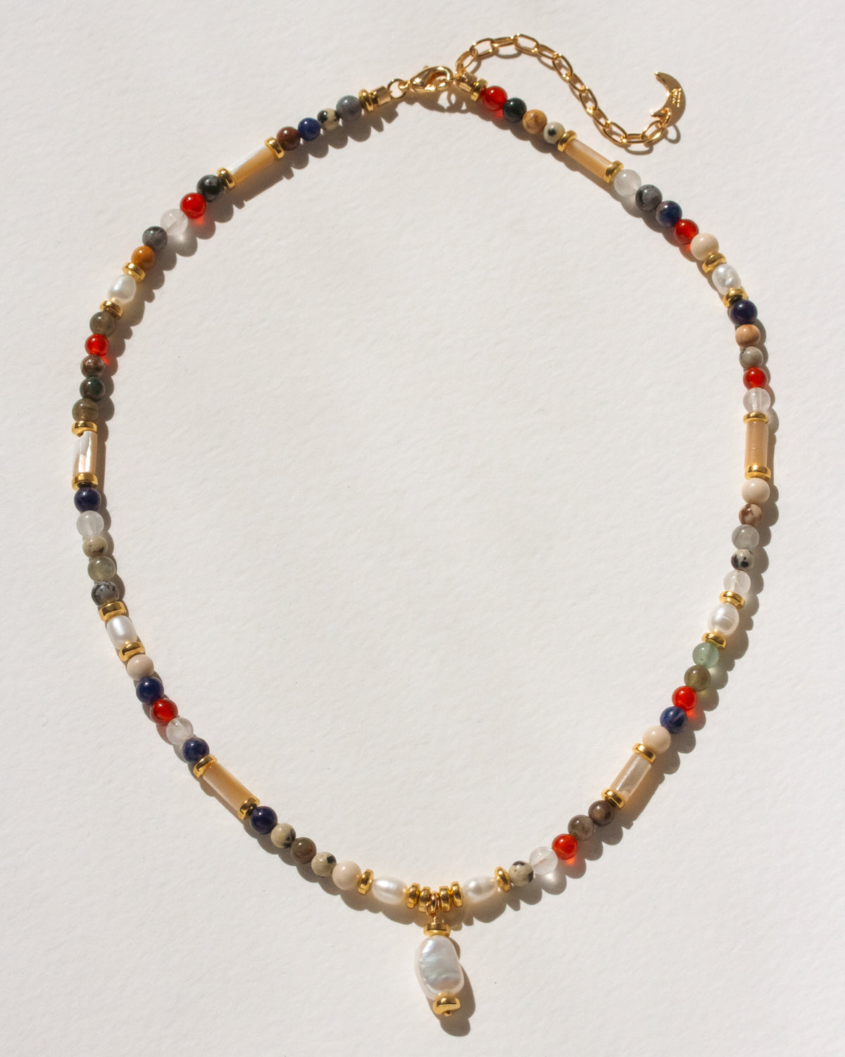 A beaded necklace with a gold clasp and a small hanging pearl charm, featuring mixed-color beads including cream, red, blue, gray, and gold-toned elements, displayed on a light off-white background.