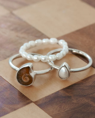 Two stacking silver rings with on a wooden surface, one with ammonite and the other with mother of pearl; and a third pearl stretch ring.