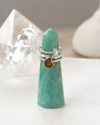 Greenish gemstone tower with a silver ring stacking set featuring a ammonite, and moonstone charms on a white surface.
