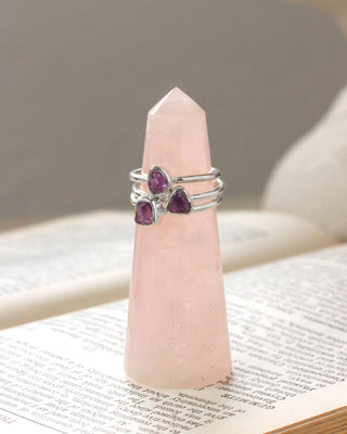 Three silver rings with purple amehtyst gemstones displayed on a pink rose quartz crystal tower sitting on an open book page.