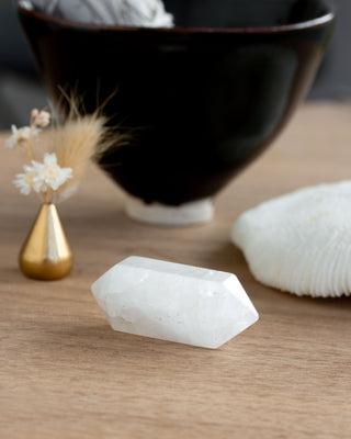 White double point quartz crystal on a wooden surface with a black bowl and decorative elements in the background