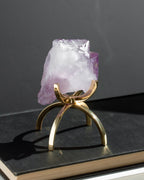 A raw amethyst crystal holder on a polished gold metal stand, displayed on a dark book against a dark gray background.