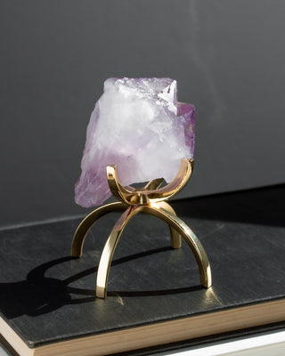 A raw amethyst crystal holder on a polished gold metal stand, displayed on a dark book against a dark gray background.