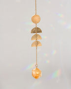 A gold-toned dangle window hanger featuring a round bead, three textured semicircle charms, and a faceted crystal pendant, suspended against a light gray background with soft rainbow light leaks.