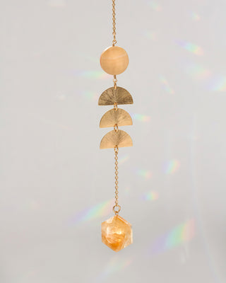 A gold-toned dangle window hanger featuring a round bead, three textured semicircle charms, and a faceted crystal pendant, suspended against a light gray background with soft rainbow light leaks.