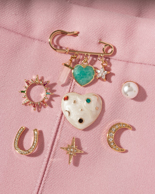 A gold-tone safety pin brooch decorated with dangling charms, including a green heart, a CZ star, and a rose quartz crystal point, displayed on a pink fabric background alongside other gold and CZ pins with celestial theme.