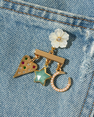 A close-up of a gold-tone charm earring with a white MOP flower, a gold heart with colorful rhinestones, a mint Amazonite star, and a gold crescent moon, resting on a blue denim background.