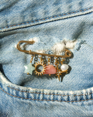A gold safety pin brooch with dangling charms, including a aqua star, a gold sun, a pink opal cloud, and a white pearl, pinned onto a blue denim jacket.
