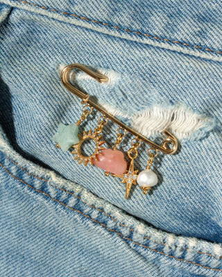 A gold safety pin brooch with dangling charms, including a light blue aquamarine star, a pink opal cloud, a gold sun, and a white pearl, pinned onto a light blue denim pocket.