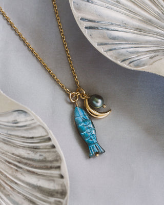 Necklace with blue labradorite fish charm and freshwater pearl, and gold crescent moon charm on a decorative surface