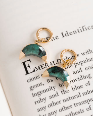 Two emerald earring charms with a crescent moon shape, attached to gold-colored hoop closures, displayed on a paper with text.