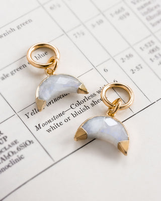 Two moonstone earrings with a crescent moon shape, attached to gold-colored hoop closures, displayed on a paper with text.
