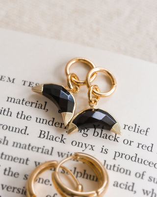Two onyx earring charms with a crescent shape, attached to gold-colored hoop closures, displayed on a paper with text.