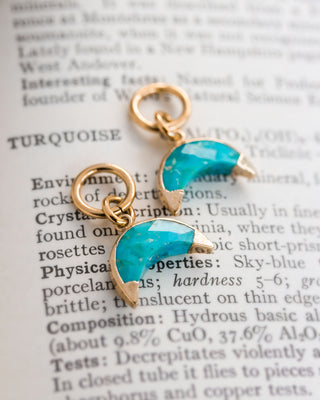 Two turquoise earring charms with a crescent shape, attached to gold-colored hoop closures, displayed on a paper with text.